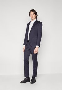 Navy suit with a single-breasted blazer and slim trousers. White dress shirt underneath. Black shoes and textured fabric details visible.