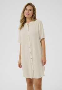 Robe beige à manches courtes en tissu texturé, avec un devant à boutons et un col rond, tombant juste au-dessus du genou.