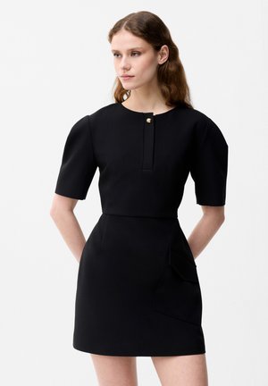 Młoda kobieta ubrana w czarną sukienkę z krótkim rękawem z bufkami na ramionach, z frontowym zapięciem na guziki i dużą boczną kieszenią, stojąca na białym tle.