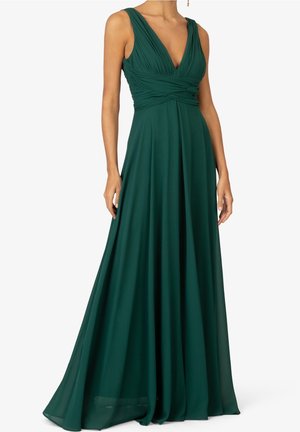 Kobieta ubrana w zieloną, bezrękawową suknię na całej długości z głębokim dekoltem w serek i marszczeniami w talii.