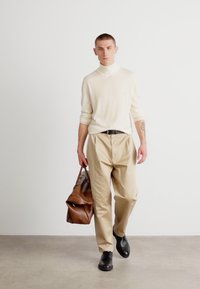 Pull à col roulé beige clair, pantalon crème, chaussures noires, tenant un sac en cuir marron, debout sur un sol gris clair devant un fond blanc.