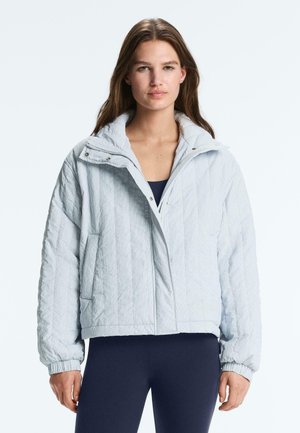 Žena oblečená v svetlomodrej prešívanej bunde so cvokmi a elastickými manžetami, cez tmavý top a nohavice, stojaca pred jednofarebným pozadím.