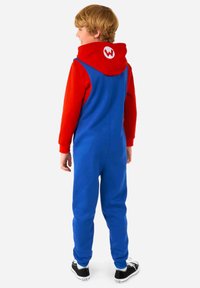 Mono con capucha azul y rojo con un gran logo blanco en forma de "W", tejido suave, corte relajado y piernas ajustadas, combinado con zapatillas negras.