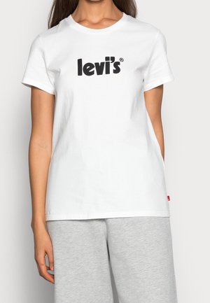 Kvinde iført en hvid Levi's T-shirt med sort logo og lysegrå bukser, vist fra nakke til hofter mod en ensfarvet baggrund.