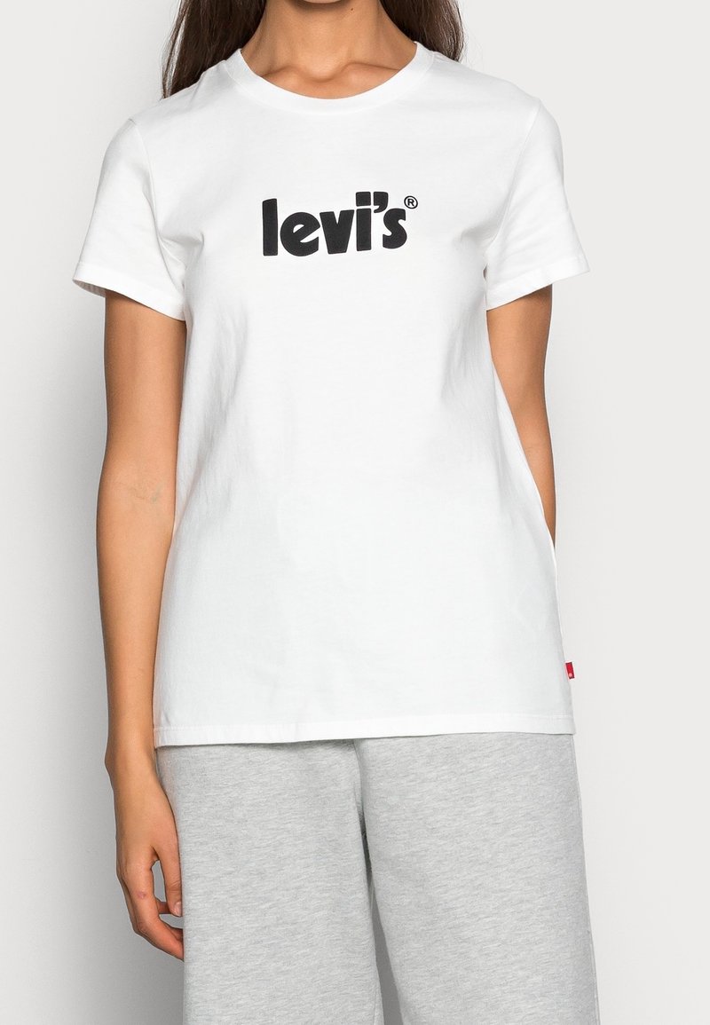 Femme portant un t-shirt blanc Levi's avec un logo noir et un pantalon gris clair, montrée du cou aux hanches sur un fond uni.