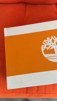 Oranžno-bež pravokotna škatla z belim logotipom drevesa na oranžni površini, postavljena na oranžno rebrasto tkanino.