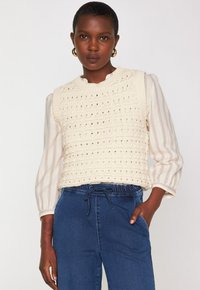 Gilet di maglia color crema con trama aperta, scollatura rotonda, indossato sopra una blusa a righe chiara con maniche a palloncino. Abbinato a pantaloni blu scuro.