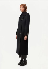 Lång svart dubbelknäppt rock med stor krage, guldknappar, sidfickor och strukturerad silhuett. Bärs över tajta svarta byxor och ankelboots.