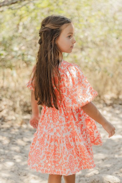 Robe blanche avec un motif floral orange, manches bouffantes courtes et volants superposés. Cheveux longs tombant sur les épaules ; debout sur un sol sablonneux.