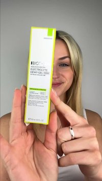 Mujer rubia sosteniendo cerca de la cámara una caja del producto de cuidado de la piel Biotherm Aquasource+ Electrolyte Dewy Gel 100H, sonriendo.