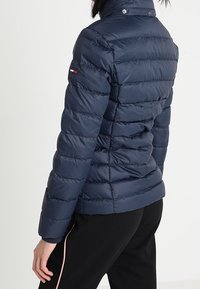 Tmavě modrá prošívaná bunda s vodorovným prošíváním, vysokým límcem a malým logem na rukávu. Hladká textura a přiléhavý střih.