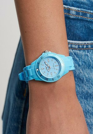 Lyseblåt armbåndsur med roterende krans og silikonerem båret på en persons håndled mod en denimbaggrund.