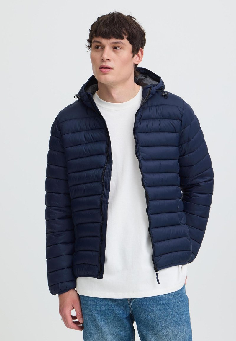 Jonge man met een marineblauwe gewatteerde jas over een wit T-shirt en blauwe jeans staat licht naar links gewend tegen een effen lichte achtergrond.