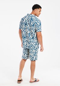 Conjunto de camisa de manga corta y pantalones cortos en blanco con un patrón de hojas en espiral en azul marino. Las telas son ligeras y informales. Chanclas visibles.