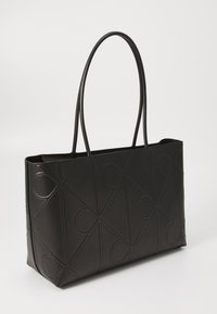 Zwarte lederen tote bag met geometrisch reliëfpatroon, verlengde handvatten, open bovenkant en een gestructureerde vorm. Glad textuur met minimale accenten.
