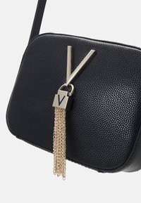Borsa nera tessuta con un accento geometrico in hardware dorato e dettaglio a frangia con catena. Chiusura con zip liscia e una singola tracolla per il trasporto.