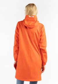 Waterdichte oranje regenjas met een capuchon, elastische manchet die kan worden aangespannen en een rechte snit. Glad textuur, middellange lengte tot halverwege de bovenbenen en zonder zichtbare sluitingen.