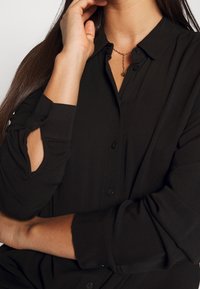 Chemise noire à boutonnière avec manches longues présentant une découpe au niveau du coude, en tissu doux, associée à un simple collier en or.