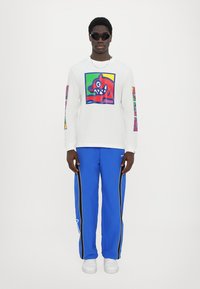 Langärmliges weißes Shirt mit buntem Grafikdruck auf der Vorderseite und den Ärmeln, kombiniert mit blauen Hosen mit seitlichen Streifen und weißen Sneakers.