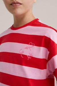 Persona que lleva una camisa con rayas horizontales rojas y rosas con pequeños símbolos rosas que incluyen un corazón, una flecha, signos de femenino y masculino en el pecho.