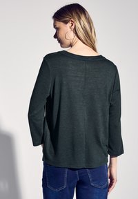 Tumma vihreä, kevyt, pitkähihainen paita, jossa on rento istuvuus ja pyöreä kaula-aukko; siinä on hienoinen tekstuuri ja normaalia matalammat olkapäät.