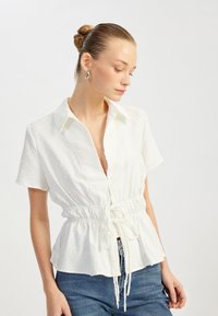 Hvid kortærmet bluse med krave, struktureret stof og rynket talje detaljer, parret med blå denim jeans.