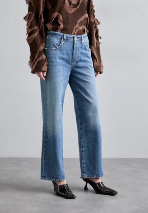 Personne portant un jean bleu clair droit, un pull marron usé et des chaussures noires à talons, debout sur un sol gris.