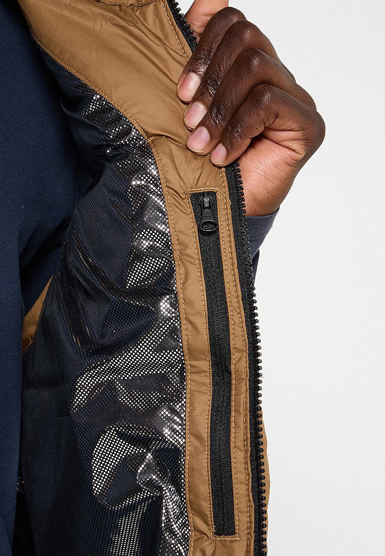 Brązowy wnętrze kurtki z teksturowanej tkaniny z błyszczącą sekcją siatkową oraz czarną kieszenią na zamek. Dłoń trzymająca okolice zamka.