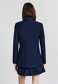 Donna vista da dietro che indossa un blazer blu scuro e una gonna plissettata abbinata su uno sfondo chiaro e semplice.