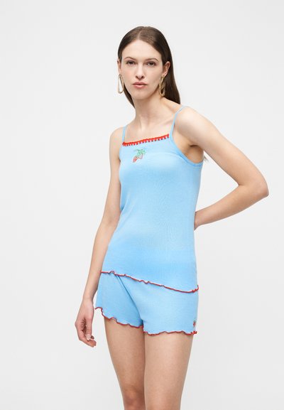 Γυναίκα που φοράει ανοιχτό μπλε αμάνικο τοπ και σορτς με κόκκινο περίγραμμα και μικρή κέντημα καρότου, στέκεται μπροστά σε απλό φόντο.