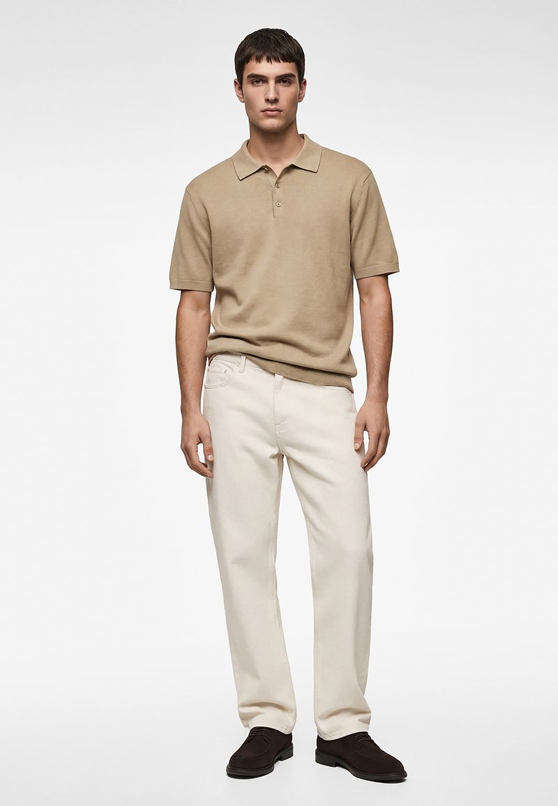 Jeune homme debout face à l'avant, portant un polo beige à manches courtes, un pantalon droit crème et des chaussures marron foncé, sur un fond uni.