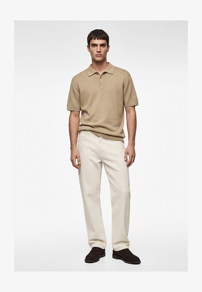 Jeune homme debout face à l'avant, portant un polo beige à manches courtes, un pantalon droit crème et des chaussures marron foncé, sur un fond uni.
