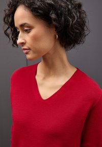 Czerwony sweter z dzianiny z dekoltem w serek, z prążkowanym wzorem. Modelka ma krótkie, kręcone włosy i stoi na szarym tle.