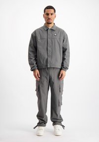 Grijze, lichte jas met knopen en een klein logo, gecombineerd met bijpassende cargobroek met meerdere zakken en elastische manchetten. Witte sneakers.