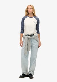 Crème lange mouwen raglan t-shirt met marineblauwe mouwen, gecombineerd met lichtblauwe jeans en een grijze gebreide trui om de taille geknoopt. Zwarte schoenen.