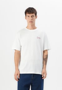 Ung mand med kort brunt hår iført en hvid t-shirt med rød tekst "Dystopia Sounds" og blå jeans, stående mod en enkel baggrund.