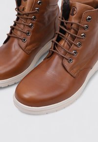 Bottes ankle en cuir marron avec des semelles en caoutchouc blanches et des œillets en métal ronds, lacées, présentées en gros plan à un angle sur un fond blanc.