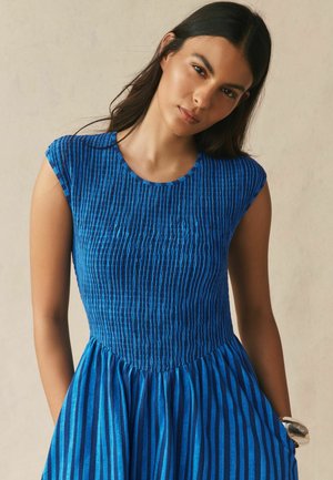 Jauna sieviete ar gariem tumšiem matiem, tērpusies zilā īsroku kleitā ar vertikālām svītrām un teksturētu augšdaļu, stāv uz neitrāla fona.