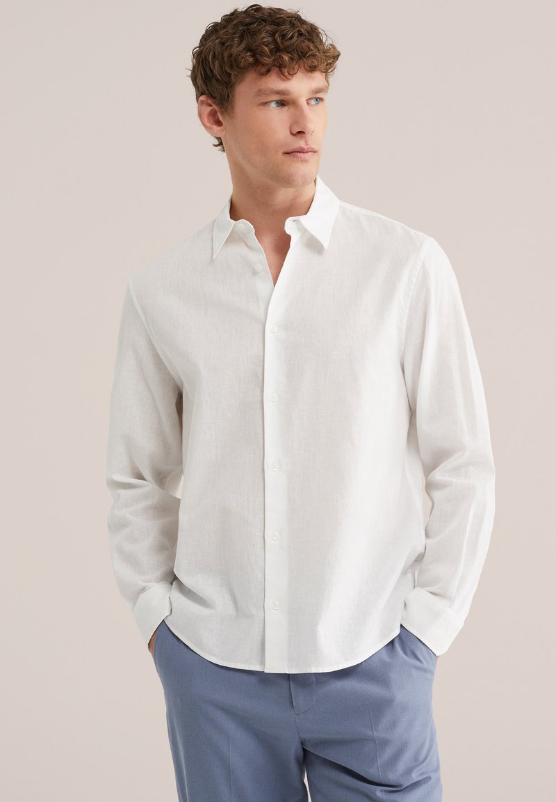 Mladý muž s kudrnatými vlasy, oblečený v bílé košili na knoflíky a modrých kalhotách, s rukama v kapsách, dívá se stranou na neutrálním pozadí.