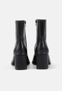 Botas de tobillo de cuero negro con un diseño elegante, tacones altos y gruesos, y cierres de cremallera en la parte posterior. Textura suave con acentos mínimos.