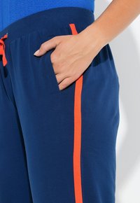 Marineblauwe joggers met een elastische tailleband en een oranje trekkoord. Voorzien van zijzakken en een oranje zijstreep over de lengte. Gladde textuur.