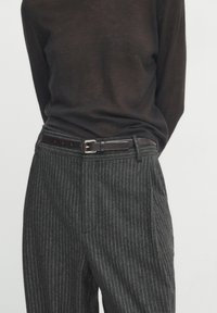 Person wearing dark brown sweater tucked into gray pinstriped trousers with a dark brown leather belt, hands behind back.