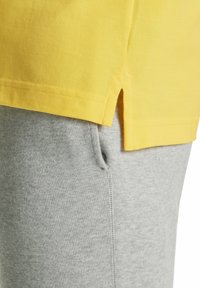 Gelb BaumwollT-Shirt mit seitlichen Schlitzen, in Schichten über hellgrauen Baumwoll-Sweatpants mit sichtbarem Taschen und glatter Textur.