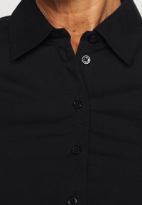 Camisa negra de botones, con cuello puntiagudo, placap de frente con seis botones negros y textura suave. Sin patrones ni acentos visibles.