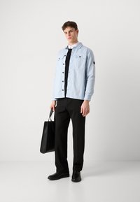 Hele sinine särk nööpidega, millel on kaks rinna-taskut, must T-särk all, mustad püksid, mustad kingad ja must kotike käes.