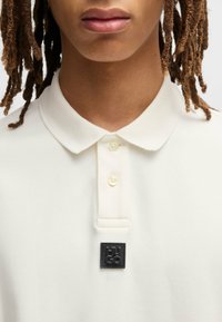 Weißes Polo-Shirt mit klassischem Kragen, drei Knöpfen und einem schwarzen HU GO-Logo-Patch auf der linken Seite. Glatte Textur und taillierte Passform.