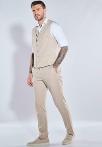 Beige Leinenanzug mit taillierter Weste, Knopfdetails und passender Hose. Darunter ein weißes Hemd. Legere Slipper. Dezente Textur.