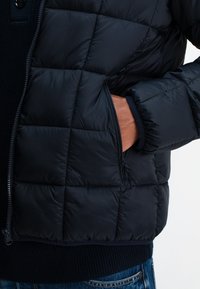 Navy Puffer-Jacke mit gestepptem Muster, ausgestattet mit einer Reißverschlusstasche und gerippten Bündchen. Das Material scheint glatt und spiegelnd zu sein.