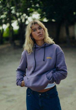 Fioletowa bluza z kapturem z czarnymi sznurkami, ozdobiona żółtym falistym logo. W połączeniu z niebieskimi dżinsami tworzy swobodny look na tle rozmytego, zewnętrznego tła.