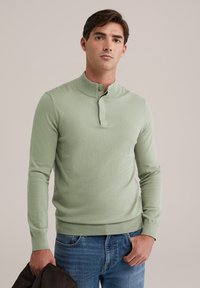 Világos zöld, hosszú ujjú kötött pulóver bordázott gallérral és gombos záródással. Az anyag lágyan néz ki, és testhezálló fazonú. Kék farmernadrágot visel.